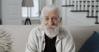 Kameraya bakıp gülümseyen yaşlı gri saçlı adamın yakın görüntüsü. Sakallı emekli erkek, rahat evdeki kanepede otururken poz veriyor. Emeklilik kavramı, insanlar ve duygular.