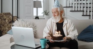 Kağıt şapkalı olgun sakallı bir adam mum üflüyor ve dizüstü bilgisayar kullanırken elinde pasta tutuyor. Yaşlı gri saçlı emekli doğum gününü internetten kutluyor. Kutlama kavramı.