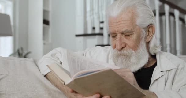 Vista de cerca de los pensionistas masculinos de pelo gris leyendo y pasando tiempo libre. Viejo barbudo disfrutando de libro mientras está sentado en un sofá en una casa acogedora. Concepto de jubilación, personas y emociones. — Vídeo de stock
