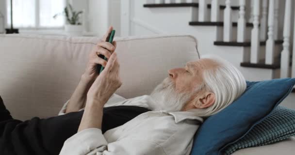 Beskär syn på mogna gråhårig man ser och vidrör telefon sreen.Äldre skäggiga manliga pensionär surfa internet och använda smartphone när du ligger på soffan hemma. Begreppet fritid. — Stockvideo