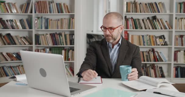 Oříznout pohled na zralého profesora v obleku a brýlích pomocí notebooku při online lekci. Muž drží šálek a mluví, zatímco pracuje u stolu. Koncepce distančního vzdělávání. — Stock video