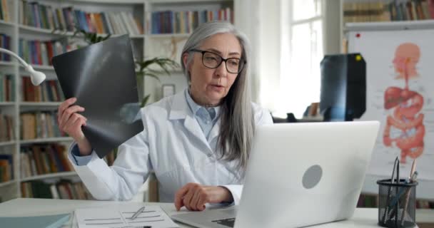 온라인 진료를 받는 동안 엑스레이 사진을 보여 주는 여성 의사의 모습. 안경을 쓰고 흰색 털을 가진 성숙 한 여성은 환자와 의사소통을 하면서 노트북을 사용 한다. — 비디오