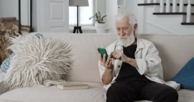 Emekli erkek emeklinin telefon içeriğini kontrol ederken ekrana bakması. Yaşlı gri saçlı adam akıllı telefon kullanırken gülümsüyor ve evdeki kanepede oturuyor..