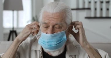 Koruyucu tıbbi maske takan ve kameraya bakan yaşlı gri saçlı emeklinin yakın görüntüsü. Evdeki koltukta oturan yaşlı adamın portresi. Koruma, virüs, hastalık kavramı..