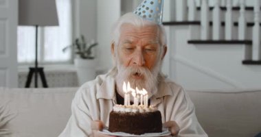 Kağıt şapkalı yaşlı sakallı adamın pasta tutarken konuşup mum üfleyişini kapatın. Gri saçlı emekli online doğum gününü kutluyor ve kanepede otururken kameraya bakıyor..