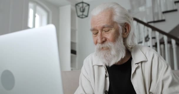 Närbild av gråhårig farfar dricka kaffe och prata medan du har videosamtal. Man pensionär med hjälp av bärbar dator för online-kommunikation medan du sitter hemma.. — Stockvideo