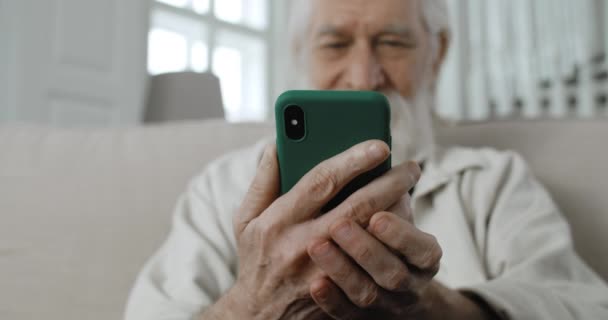 Närbild av äldre manliga pensionär chatta med familjen när du skriver på telefon sreen.Positiv skäggig man med grått hår läsa meddelande och ler när du sitter på soffan hemma. — Stockvideo