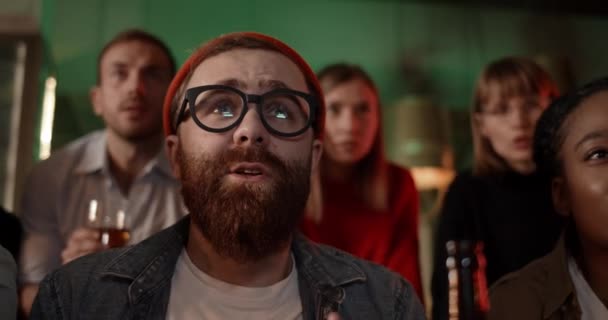 Close up view of bearded guy in glasses watching sports game with friends in pub and looking upset. Group of multiethnic people become sad while team they supported lose. — Stock Video