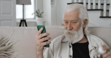 Yaşlı adamın konuşmasını ve videoya çekilirken şişeyi haplarla göstermesini yakından izleyin. Sakallı emekli erkek, akıllı telefon kullanarak internette sohbet ediyor ve evdeki kanepede oturuyor..
