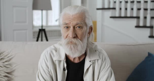 Vista de cerca del hombre de pelo gris senior en los años 70 girando la cabeza y mirando a la cámara. Retrato del abuelo barbudo posando sentado en un sofá en una casa elegante. Concepto de jubilación. — Vídeo de stock