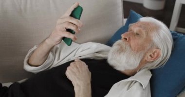 Yaşlı gri saçlı dedemin telefon kulübesine baktığı mahsul manzarası. Sakallı, yaşlı bir erkek emeklinin akıllı telefon kullanırken video izlemesi ve evdeki kanepede yatması. Boş zaman kavramı..