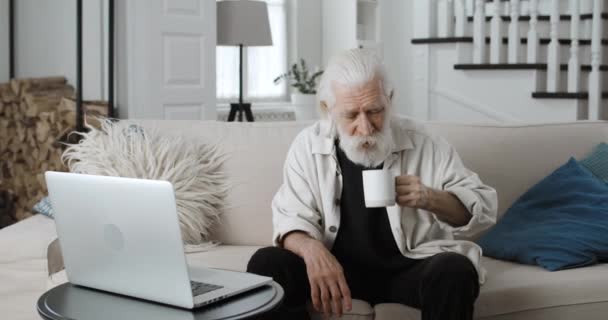 Crop view van senior grijze harige man praten en het drinken van koffie tijdens het hebben van videogesprek. Man gepensioneerde met behulp van laptop voor online communicatie tijdens het zitten op de bank thuis. — Stockvideo