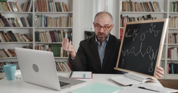 안경을 쓴 남자는 칠판에 분필로 분필을 쓴 채 노트북을 보고 있습니다. 온라인으로 자료를 설명하는 성숙 한 선생님. 교수가 말하는 것도 그렇고. 거리 학습에 대한 개념. — 비디오