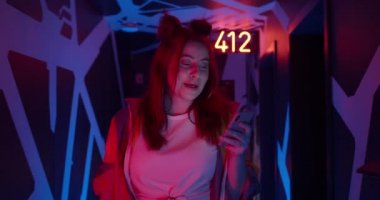 Sosyal medyada gülümseyen ve haber yayını yapan şık bir kadın görüntüsü. Koridorda neon ışıklarıyla yürürken akıllı telefonunu kullanan modaya uygun kıyafetli çekici bir kadın..