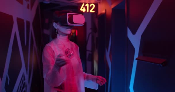 Persona femenina con gafas VR futuristas y ropa de moda de pie en la habitación con iluminación de neón. Mujer en ropa de moda moviendo las manos y divirtiéndose mientras experimenta la realidad virtual. — Vídeos de Stock
