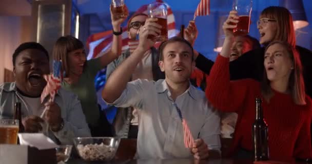 Glada multietniska människor med amerikanska flaggor rakning och klinkande öl samtidigt stödja landslaget. Grupp av olika vänner firar och ler medan du tittar på sport spel på puben. — Stockvideo