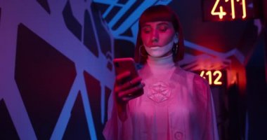 Bilimkurgu dünyasındaki kadın arka planda parlak sayılar ve neon ışıklarla yürürken akıllı telefon kullanıyor. Burun halkalı bir kadın gelecekteki oteldeki telefon ekranına bakıyor..