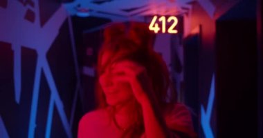 Genç ve çekici bir bayanın iyi ruh hali içinde yürümesini ve kameraya bakmasını sağlayın. Kameraya poz veren ve neon ışıklara gülümseyen neşeli bin yıllık kadın model. Kapalı.