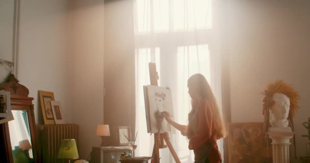 Lviv, Ukraine - 17 janvier 2020 : Femme peignant au pinceau sur papier à la lumière du soleil debout devant un chevalet. Artiste féminine dans le processus de création d'image à l'atelier d'art confortable. — Video