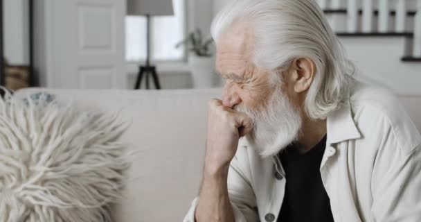 Крупним планом вид розчарованого старого, який думає, піднімаючи голову. Бородатий чоловік пенсіонер виглядає засмученим, сидячи на дивані в затишному будинку. Концепція виходу на пенсію, люди та емоції . — стокове відео