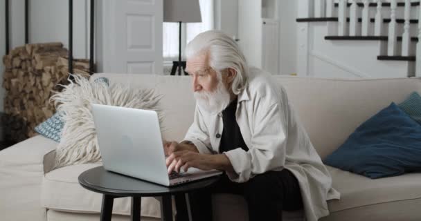 Widok na dojrzałego siwego mężczyznę piszącego na maszynie podczas komunikacji online. Mężczyzna brodaty emeryt za pomocą laptopa podczas siedzenia na kanapie w domu. Pojęcie czasu wolnego i emerytury. — Wideo stockowe