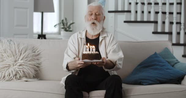 Άποψη των ώριμων γενειοφόρος άνθρωπος σε χάρτινο καπέλο φυσώντας κεριά, ενώ κρατώντας κέικ. Senior γκρίζα μαλλιά συνταξιούχος γιορτάζει τα γενέθλια σε απευθείας σύνδεση και ψάχνει για κάμερα.Έννοια της γιορτής. — Αρχείο Βίντεο