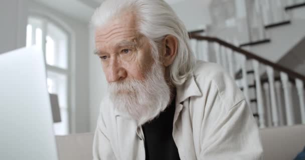 Vista de perto do avô grisalho maduro digitando enquanto conversa online. Homem aposentado barbudo usando laptop enquanto sentado no sofá em casa. Conceito de lazer e aposentadoria. — Vídeo de Stock