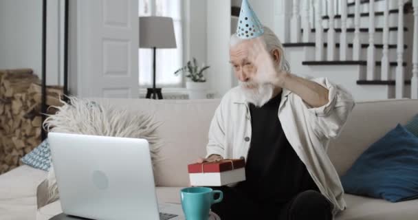 Dojrzały brodaty mężczyzna w papierowej czapce dmuchając świece i trzymając ciasto podczas korzystania z laptopa. Starszy siwy emeryt świętujący urodziny online. Pojęcie uroczystości. — Wideo stockowe