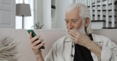 Video görüşmesini bitirirken el sallayan ve öpüşen yaşlı gri saçlı adamın yakın görüntüsü. Evde internet üzerinden iletişim kurarken gülümseyen ve akıllı telefon kullanan pozitif sakallı emekli..