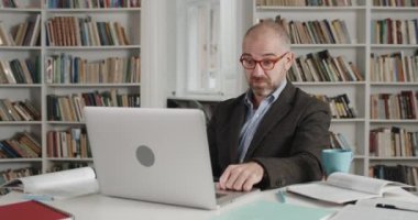 Dizüstü bilgisayar kamerasıyla konuşan gözlüklü olgun bir adamın görüntüsü. Takım elbiseli bir erkek öğretmen kitap dolu bir odada otururken online ders alıyor. Eğitim kavramı, uzaklık öğrenimi.