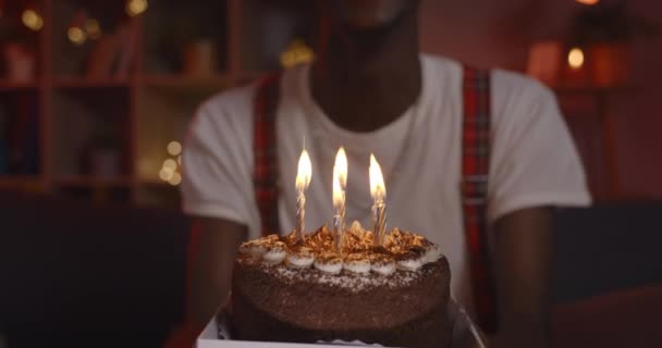 Crop άποψη της Αφρικής Αμερικανός αρσενικό πρόσωπο στο καπέλο γενεθλίων χαρά και να μιλάμε, ενώ κοιτάζοντας προς την κάμερα. Θετικός άντρας με βαμμένα μαλλιά που φυσάει κεριά στην τούρτα ενώ κάθεται στον καναπέ. — Αρχείο Βίντεο