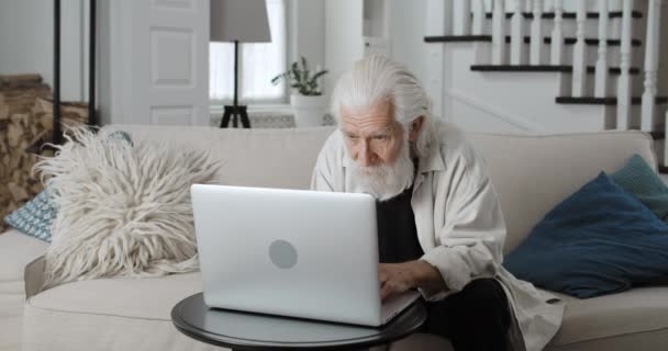 Crop view of senior grey haired man typing while browsing internet. Male bearded retiree using laptop while sitting on sofa at home. Concept of leisure and retirement,modern technology. — Stock Video