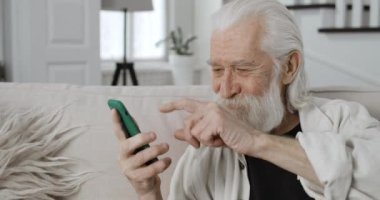 İnternette gezinirken neşeli erkek emeklilerin ekili görüntüsü kayıyor ve telefon ekranına bakıyor. Yaşlı sakallı adam akıllı telefon kullanarak gülümsüyor ve evdeki kanepede oturuyor..
