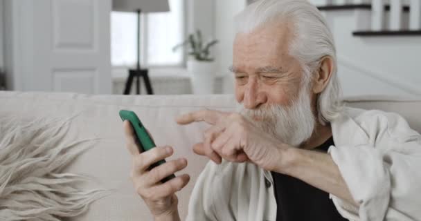 在浏览互联网的时候，可以看到快乐的男性养恤金领取者滚动和看手机屏幕的照片。老头儿笑着用智能手机，坐在家里的沙发上. — 图库视频影像