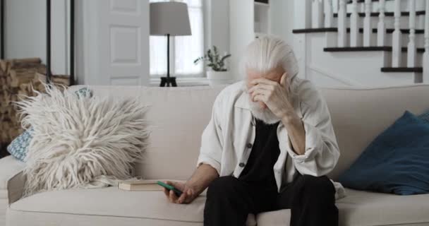 Вид на сумного старого, який піднімає голову і тримає смартфон. Сіра волохата чоловіча пенсіонерка почувається самотньо, сидячи на дивані вдома. Концепція поганих новин, самотності . — стокове відео