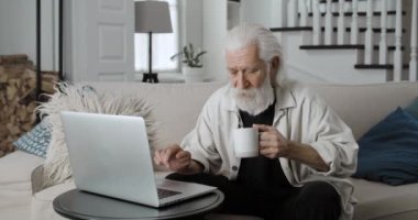 Erişkin gri saçlı bir adamın internette gezinip kahve içtiği ekin manzarası. Erkek sakallı emekli evdeki kanepede otururken dizüstü bilgisayar kullanıyor. Boş zaman ve emeklilik kavramı.