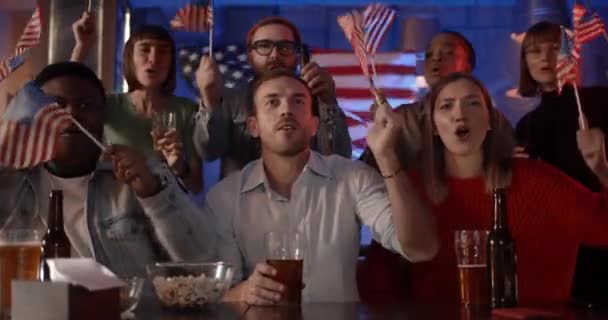 Pessoas divertidas alegres acenando bandeiras americanas e cantando enquanto apoiam a equipe nacional. Pessoas multiétnicas felizes se alegrando e sorrindo enquanto assiste sportrs jogo com cerveja no pub. — Vídeo de Stock