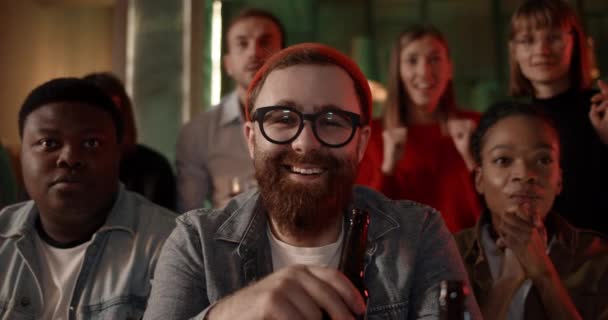 Homme barbu dans des lunettes et chapeau tenant bouteille de verre de bière tout en regardant à la caméra et rire. Des gens joyeux et diversifiés se réjouissant et célébrant tout en regardant le sport dans un pub. — Video