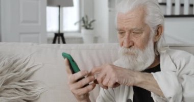 Gri saçlı emeklinin ekili görüntüsü kayıyor ve telefon ekranına bakıyor. Yaşlı sakallı adam internette gezinirken akıllı telefon kullanıyor ve evdeki kanepede oturuyor..