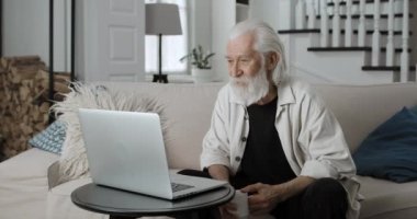 Yaşlı, kır saçlı adamın video izleyip çay içtiği bir manzara. Erkek sakallı emekli evdeki kanepede otururken dizüstü bilgisayar kullanıyor. Boş zaman, eğlence ve emeklilik kavramı.