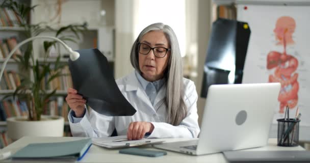 Médica feminina segurando a foto de raio-x do peito ao ter consulta médica on-line no escritório. Mulher idosa em óculos e assaltos brancos usando laptop enquanto se comunica com paciente. — Vídeo de Stock