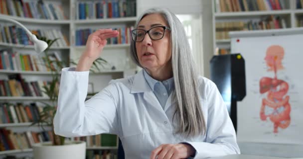 Femme médecin mature dans des lunettes montrant au patient comment respirer correctement tout en ayant une consultation vidéo. Femme en blanc voler parler et gesticuler tout en étant assis dans le bureau médical. — Video