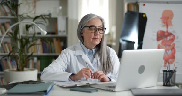 Moden kvindelig læge i briller, der har videoopkald, mens du sidder ved bordet i kontoret. Ældre kvinde i hvid medicinsk røve taler og smiler, mens de har konsultation med patienten online. – Stock-video