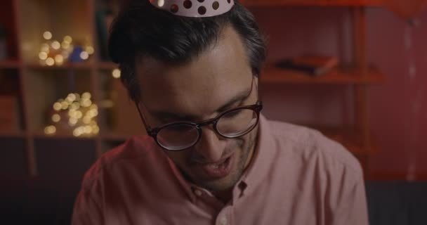 Doğum gününü kutlarken kameraya bakan neşeli adamın yakın görüntüsü. Kağıt şapkalı mutlu bir erkek hediye kutusunu açarken seviniyor ve "Vay canına" diyor.. — Stok video