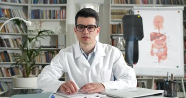 Gözlüklü profesyonel bir adam video sohbeti yaparken hastanın tıbbi formunu dolduruyor. Beyaz soygunu yapan erkek doktor konuşuyor, yazıyor ve sağlık ofisinde kameraya bakıyor..