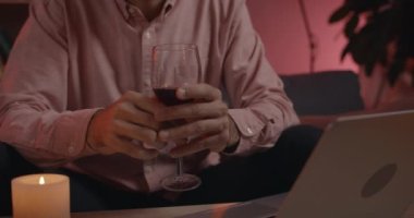 Çevrimiçi video konuşması yaparken şarap kadehi tutan erkeklerin el görüntüsü. Kimliği belirsiz bir adam evdeki koltukta otururken bilgisayar ekranına bakıyor..