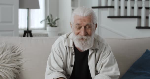 カメラを見ながら話す先輩ひげを生やした男のミステリービュー.自宅でソファに座っている間、ビデオ通話を持つ陽気な灰色の髪の男性の退職者.オンラインコミュニケーションの概念. — ストック動画