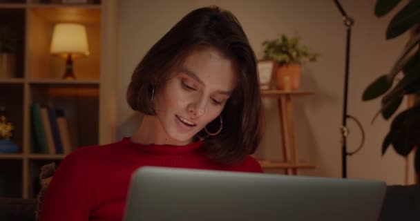 Närbild av tusenårig kvinna som använder laptop när du sitter på soffan hemma. Ganska brunett kvinnlig person som surfar på internet och ler. Begreppet fritid och fritid. — Stockvideo