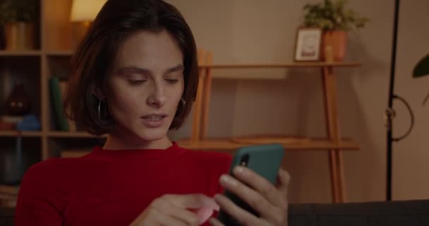 Zamknij widok atrakcyjnej kobiety przewijania ekranu telefonu i mówiąc wow siedząc na kanapie w domu. Brunetka młoda kobieta za pomocą smartfona i patrząc zaskoczony. — Wideo stockowe