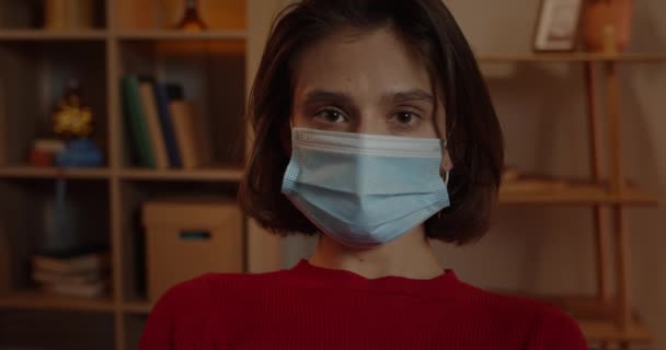 Portrait of young woman in medical protective mask looking to camera. Close up view of millennial female person with short brunette hair sitting at home. Concept of quarantine, viruses. — Stock Video